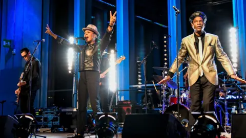 The Selecter performing on stage. The lead singer, called Pauline Black, is making the international peace symbol with both hands.