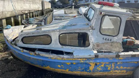 A derelict looking small boat in a harbour while the tide is out. It is white and blue with patches of badly flaking yellow paint on the hull. and a life ring on its roof.
