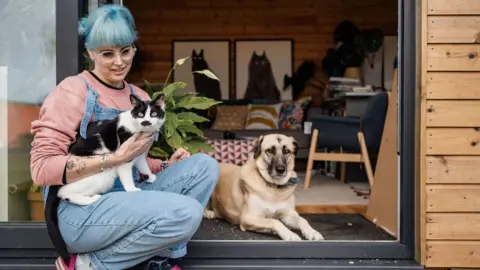 Harriet Lowther A woman with grey-blue hair, glasses and wearing denim dungarees sat in the open door way of her art studio holding a black and white cat, with a large beige-coloured dog lying down next to her - with pieces of art of cats on the wall in the background. 