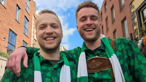 Two men, smiling at the camera, standing in a street. They are both wearing a green shamrock-covered shirt, with a scarf in the colours of the Irish flag - green, white and gold. One of them is wearing lederhosen or traditional leather breeches worn in Germany. 