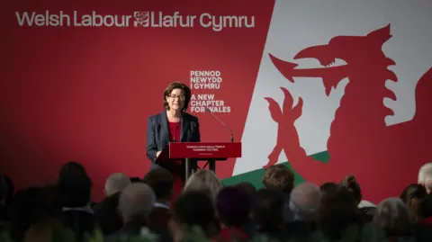 Getty Images A woman, Eluned Morgan, is standing at a lectern while making a speech to an audience, and she is standing in front of a large graphic depicting a red Welsh dragon and the words "Welsh Labour, Llafur Cymru" on it.