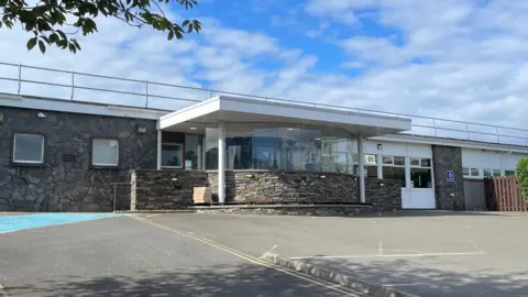 The front of a long single storey stone-clad building with a semi-circular foyer in the centre with the upper half all glass windows. There is a cloudy blue sky and the empty carpark in the foreground.