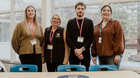 BILTT/Grange Academy Three people, all in a row, looking at the camera, all with lanyards round their necks. The woman on the left has long blond hair, is wearing a brown top and black trousers. The woman next to her has a black gilet on, brown jumper, and dark trousers. Toby has his hands crossed in front of him and is wearing a black top and black trousers. The woman to the right, has long dark hair tied back, is wearing a brown top and blue jeans. A table and three blue chairs are in front of them and they are by a large window. 