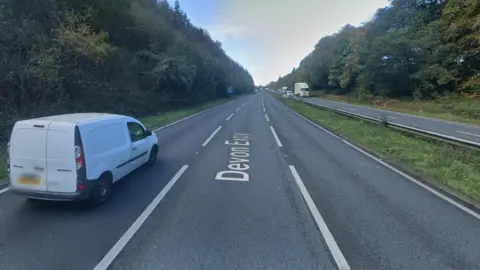 A white van drives along the A38 near Kenn in Devon on a sunny day. There are three lanes on the carriageway, which has a row of trees lining the road. On the opposite carriageway, several vehicles including a couple of lorries are driving along the two carriageways.