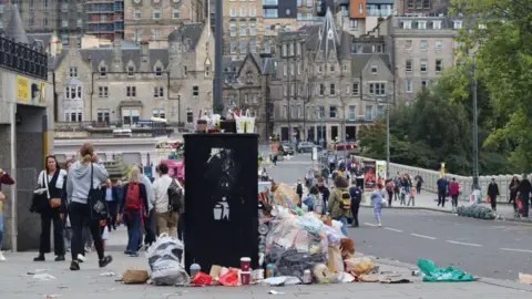 An overflowing bin is in the foreground of an Edinburgh street. There are large buildings in the background and people walking down the street. Green trees line the right of the image.