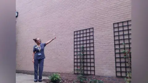 A woman in a blue uniform stands in front of a brick wall and points at it. She has her hair tied in a bun and glasses.