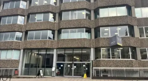 Front of an office building. It is brown coloured with large windows taking up most of the front aspect. The reception area is visible as well as the street outside. 