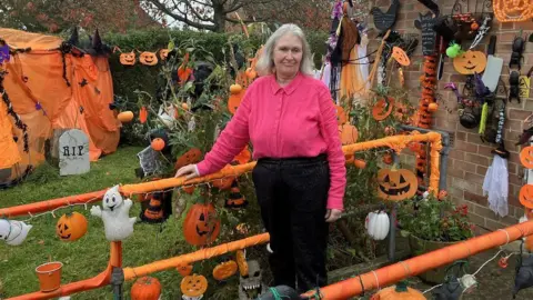 A woman who has decorated her house for Halloween for 40 years stands in her garden, surrounded by orange decorations and pumpkins. 