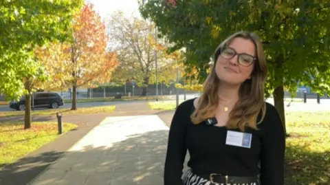 Sienna smiles at the camera. She wears glasses, has long brown-blonde hair, and has a white business card on her black top. 