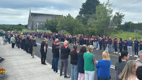People were lined up on a street outside a church. There is a cloudy sky above.