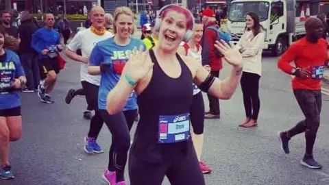 Stevie O'Brien A woman in black Lycra waves to the crowd as she runs in an organised race. Other men and women run past in the background.