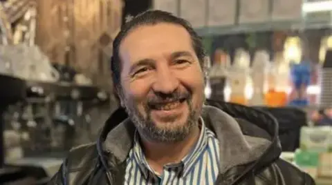 A man in a coat. He is smiling behind a bar. 