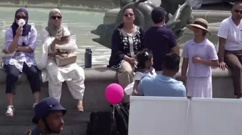 PA Media people sit on the edge of the fountain to cool off, some holding fans