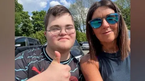 Family A boy in a black and blue stripy t-shirt with glasses and his thumb up next to a woman in a dark blue top and reflective sunglasses