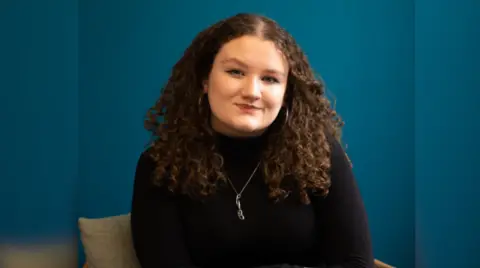 DKMS Bronagh Hughes smiles at the camera. She has thick, curly dark brown hair, is wearing hooped earrings and a high-necked black jumper, and a pendant on a thin gold chain.