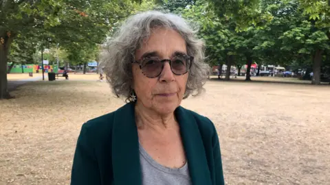 Teresa with grey curly hair, glasses and a dark green jacket stands on parched grass in a park, with trees and shops behind her.
