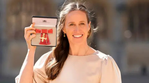 Helen Glover holding up her OBE medal and smiling. She is wearing a cream dress.