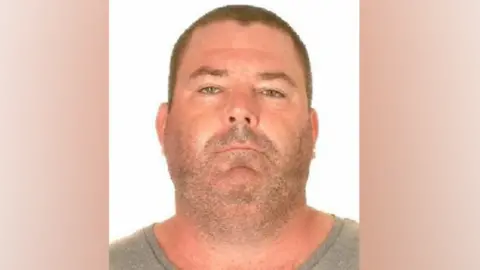 A police mugshot of a man wearing a grey t-shirt. He has very close-cropped short brown hair and stubble. He has a chubby face and neck of roughly the same width and appears to be sunburnt. 