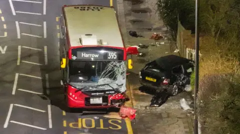 A 395 single decker bus with a smashed window and a black car which has crashed into a hedge 