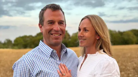 i5 Media/PA Media Peter Phillips wheres a checkered shirt and Harriet Sperling in a white blouse puts her hand with engagement ring on her fiance's chest. They stand in a field.