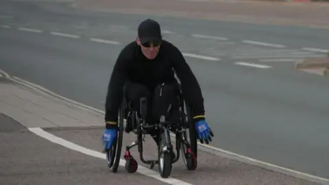 Wheelchair athlete Lexi Chambers training for her challenge along the seafront in Exmouth. She is wearing an all black outfit, with black glasses and a black cap. She is wearing blue gloves. 