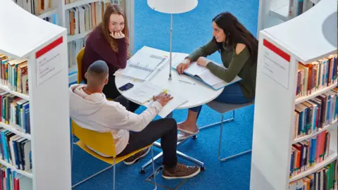 Getty Images A diverse group of students sit at a table in a university library.