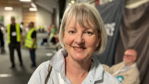 Liz Leffman, wearing a grey and white shirt with the collar turned up, smiling for the camera.