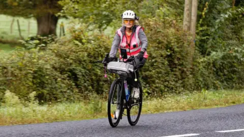 Family handout Alison Warner riding a bicyle during her cycle challenge. She has blonde hair that is pulled into a ponytail. She is wearing a white helmet and a pink high-vis jacket. She is cycling down a road, a field can be seen to the side.