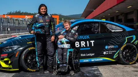 Two people in Team Brit race suits in front of a BMW M4 GT4 Race Car. It is black with blue and yellow accents. They are at the Circuit de Spa-Francorchamps in Belgium.
