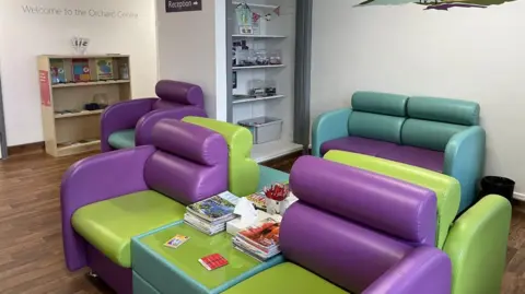 Orchard Centre Guernsey The waiting room in the Orchard Centre has five green and purple armchairs and a sofa along with a table with magazines and pens. There are two bookshelves lining the walls and writing on the wall that reads 'Welcome to the Orchard Centre'.