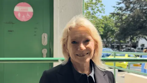 BBC Senator Elaine Millar wearing a dark jacket, standing outside a public toilet entrance, with a green accessible restroom door visible in the background.