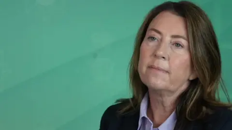 Det Supt Rachel Wilson with long brown hair wearing a lilac shirt and black blazer. She is sitting in front of a green background. She looks impassively.
