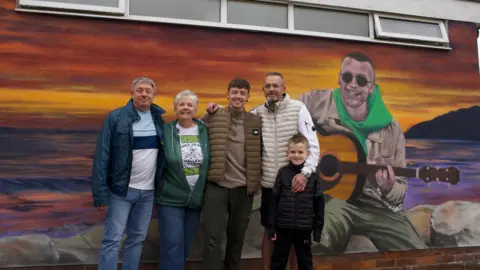 A mural of a man sat on rocks, with the sunset behind, playing a guitar. Stood in front are three men, a boy and a woman. The man on the left has grey hair, blue coat and blue jeans with a blue and white top. Next to him is a woman with grey hair, green coat and white t-shirt with blue jeans. Next to her a man with a khaki gillet and brown top and dark trousers. Next to him a man in glases with a crean gillet and white top. He is wearing glasses and in front of him is a young boy in a black top