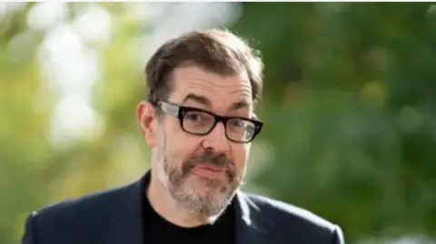 Getty A picture of Richard Osman. He is wearing a navy suit with a black t-shirt and rectangular glasses with thick, black rims. He has dark hair and a grey beard. The background is out of focus trees.