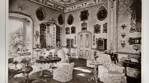 Private Collection An old, black and white photo of an ornate, vintage sitting room filled with patterned armchairs and small tables, decorated with mirrors, framed portraits, and detailed wall designs, with a large window letting in light.