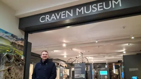 Ben Crick Ben Crick wearing a dark coat and smiling underneath a sign saying "Craven Museum" and there are objects behind him, such as a bike and signs giving information about the objects there