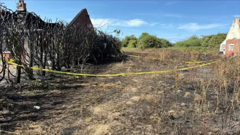 Norfolk Fire and Rescue Service Singed and charred landscape after a bonfire spreads to a neighbouring bungalow. There is yellow tape to cordon off the area affected. The sky is blue with little cloud in it.