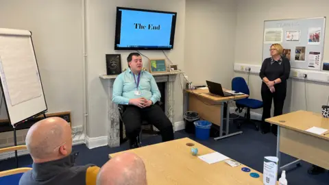 George is sitting in the front of a classroom speaking to a group of people. Behind him is a screen with a slide saying The End.