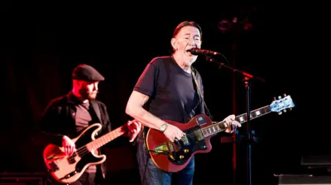 Getty/Frank Hoensch/Redferns British singer Chris Rea stands at the mic, holding a guitar. He is in the middle of singing in this picture. He has black hair slicked back and white stubble. .He wears a black t-shirt.  The picture was taken during a concert at the Tempodrom on October 30, 2017 in Berlin, Germany.