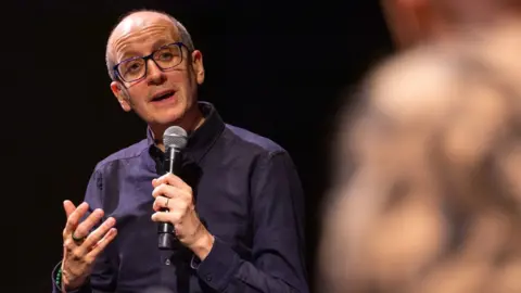 Netflix Jack Thorne speaks microphone on panel events in a dark blue shirt to promote puberty