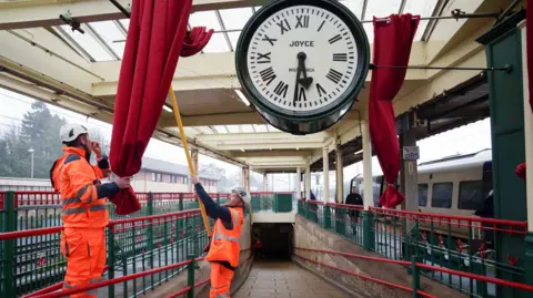 Network Rail Two workers in orange high-visibility clothing pull back red curtains to reveal the restored clock hanging above the station platform. The clock face displays the name 'Joyce' and 'Whitchurch' in the centre. A Northern train is visible on the platform to the right, with the station's distinctive green and red ironwork railings below.