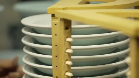 White plates are stacked up behind a yellow structure on the image. A hand is visible on the bottom left and the background is blurred.