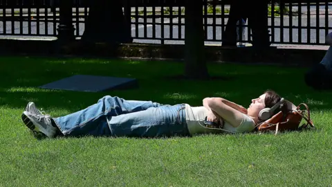 Pacemaker A woman wearing headphones lying on grass with her arms crossed and eyes closed. She has dark hair, wearing a white t-shirt, jeans and trainers.