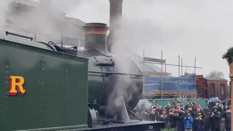 A crowd of people filming a steam locomotive on their phones and cameras as it sounds its whistle