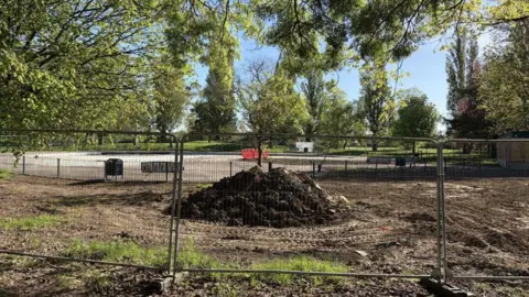 Richard Madden/BBC A mound of soil and rubble stands in the centre of a muddy clearing. It is surrounded by metal safety fencing with trees, park benches, and a paved path visible in the background.