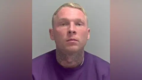 Hertfordshire Police A mug shot of Myles Judd police released in connection with an armed robbery in St Albans. He has a tattoo around his neck and is wearing a purple jumper.