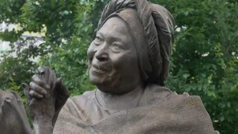 The photo is a close up of a bronze statue, it depicts a smiling woman wearing a headscarf holding hands with another woman. 