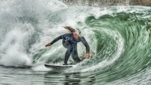 Tony Marsh A female surfer wearing a black wetsuit, emerged in a green wave. The water is splashing all around her.