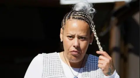 Athena Picture Agency A head and shoulders shot of Gonzales. She is holding one of her black and blonde braids in one hand. The rest of her hair is braided and tied back in a bun, and she is wearing a white long-sleeved shirt with a houndstooth pattern dress over top 