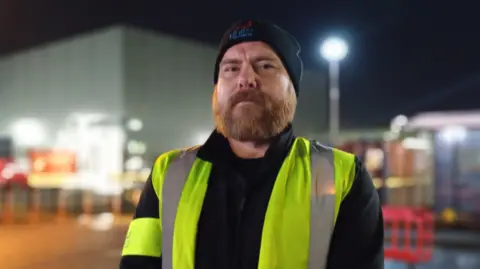 Billy looking at the camera. He has a beard and is wearing a beanie hat and high visibility vest over a black jacket. He is standing outside.